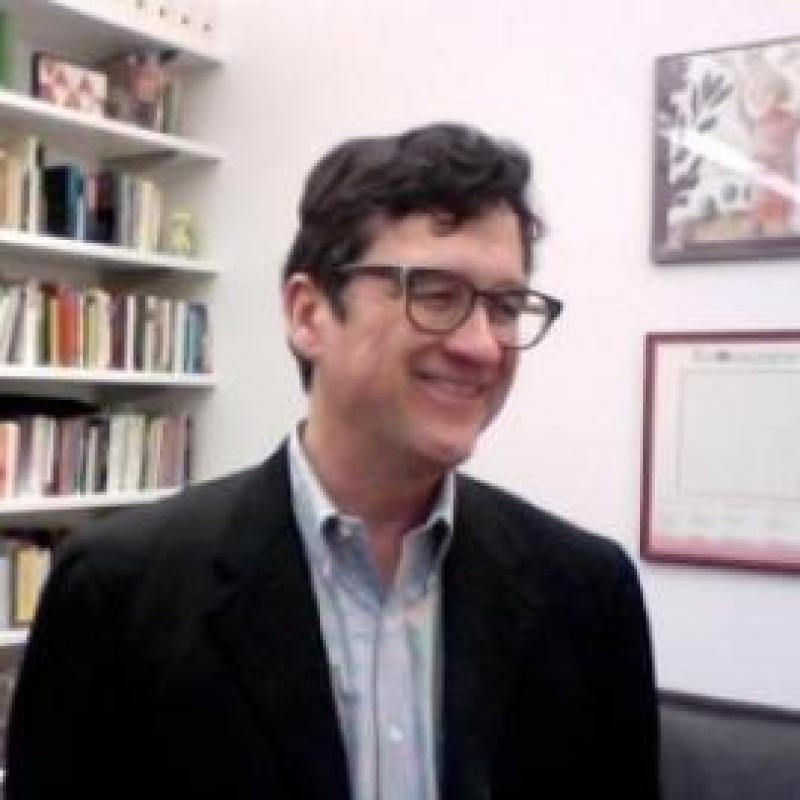 Roland Greene in a black suit with glasses standing in front of a bookcase.