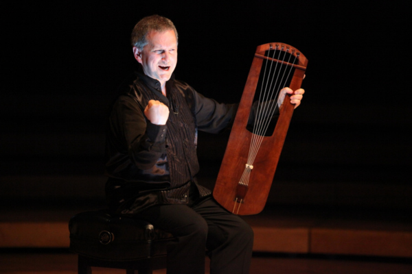 A Caucasian man on a dark stage, holding a medieval rectangular harp with his hand in a fist and his mouth open. 