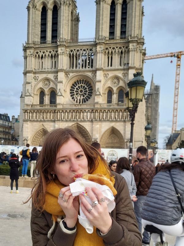 A woman with long blond hair is taking a bite out of bread in front of a gothic cathedral